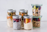 Six glass jars of make-ahead food on a white counter — chia pudding with berries, granola with fruit, oats and rice