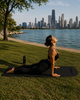 A woman in a black sports bra and leggings does a cobra pose on a black yoga mat outdoors by the water, with a city skyline in the background. A black water bottle sits beside the mat.
