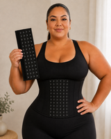 A woman wearing a black waist trainer corset holds up a matching black grommet panel in her right hand, standing in a brightly lit indoor setting.